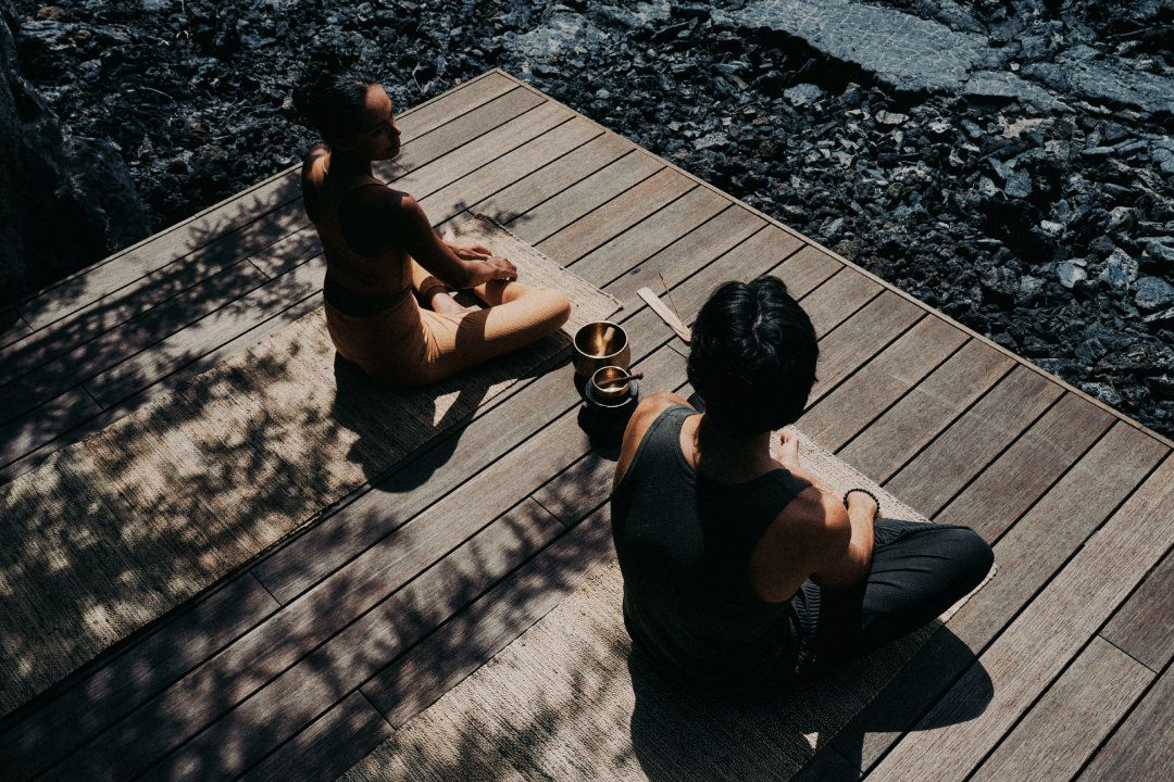 A couple sitting on a wooden deck in a tropical setting, ready for meditation.